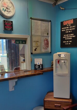 A hotel reception area with a blue wall, a clock, a welcome sign, a notice board, a water dispenser, and a date display. - Hollywood Motel (Crossett)