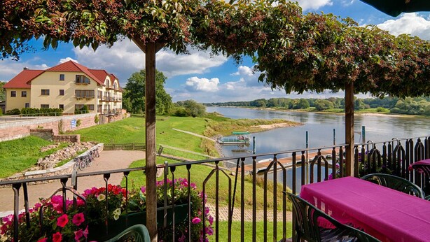 Terraza a la orilla del río con una mesa preparada para comer, con vista al río y un edificio de techo rojo.