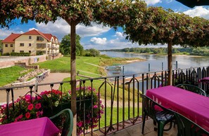 Een terras aan de rivier met een eettafel, met uitzicht op de rivier en een gebouw met een rood dak.