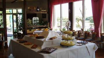 A buffet table with various desserts and a three-tiered cake, set in a room with large windows and red curtains.