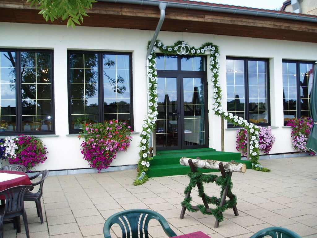 Un bâtiment à la façade blanche, doté de grandes fenêtres et d’une entrée verte. Des jardinières remplies de fleurs sont posées sur les rebords des fenêtres, et un banc se trouve à l’extérieur.