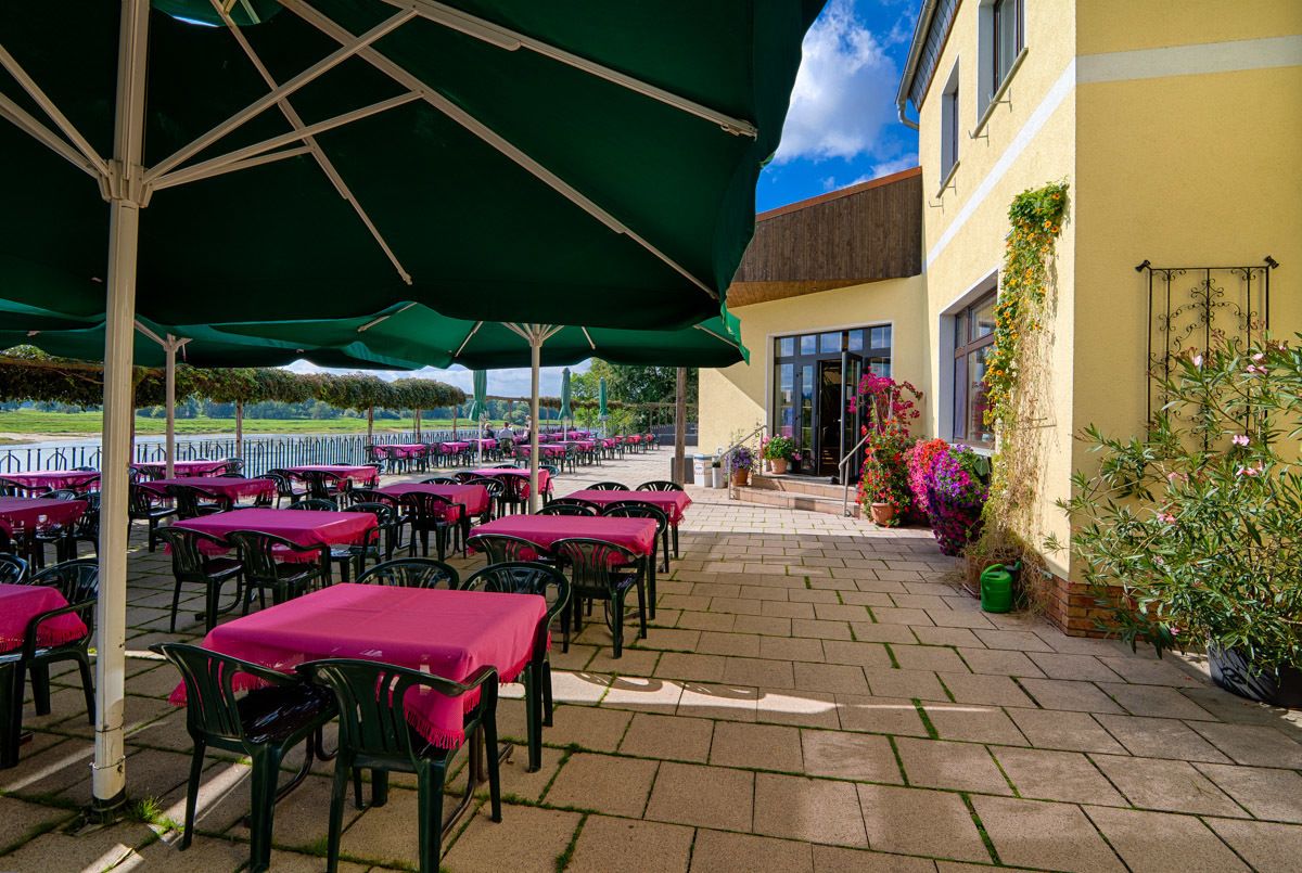 Eine Terrassengaststätte mit Tischen und Stühlen, beschattet von grünen Sonnenschirmen, und ein Gebäude mit gelber Fassade und Fenstern.