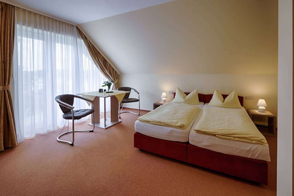Habitación de hotel con cama, dos mesitas de noche, un escritorio y una silla, y un ventanal con cortinas.