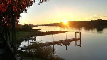 A serene lakeside view with a wooden dock extending into calm waters, surrounded by lush greenery and a clear sky.