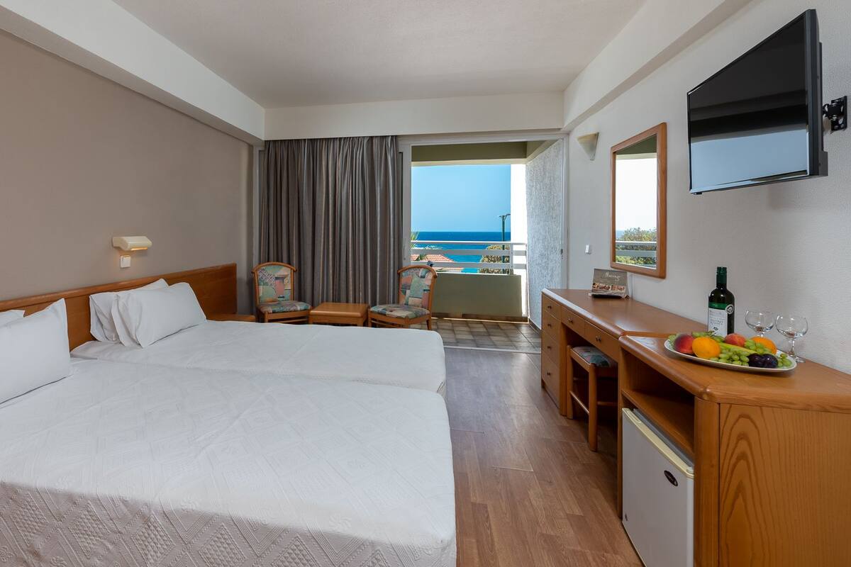 A hotel room with two beds, a desk with a fruit plate, a bottle of wine, and a TV on the wall.