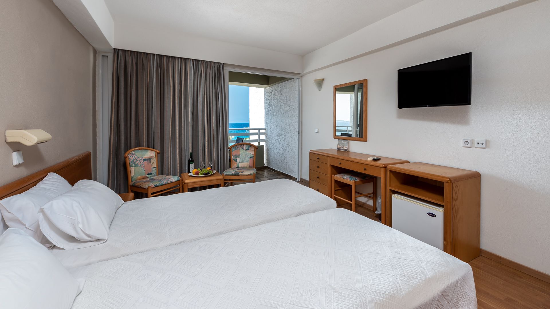 A hotel room with a bed, a desk, a chair, a dresser, a television, and a view of the ocean.