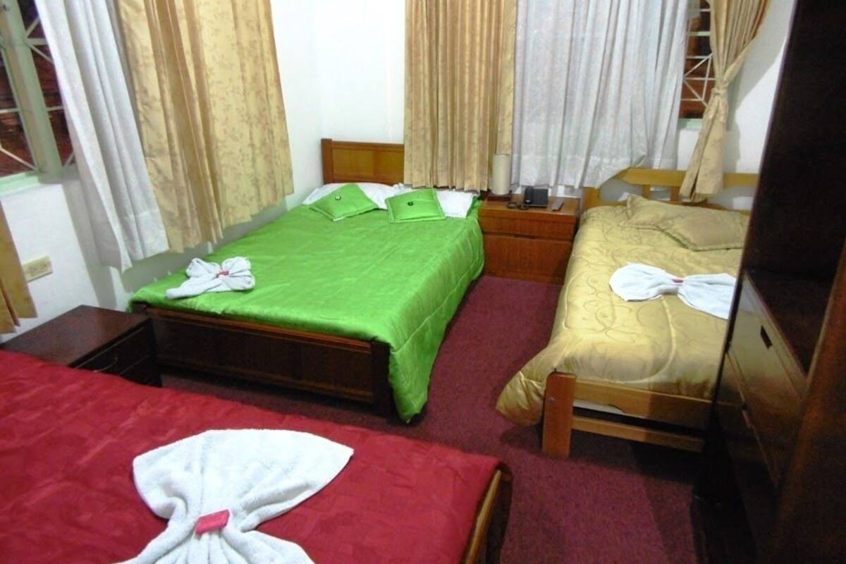 A hotel room with two single beds, each with a green and a yellow bedspread, and a red bedspread in the foreground. There is a wooden nightstand with a lamp and a telephone between the beds.