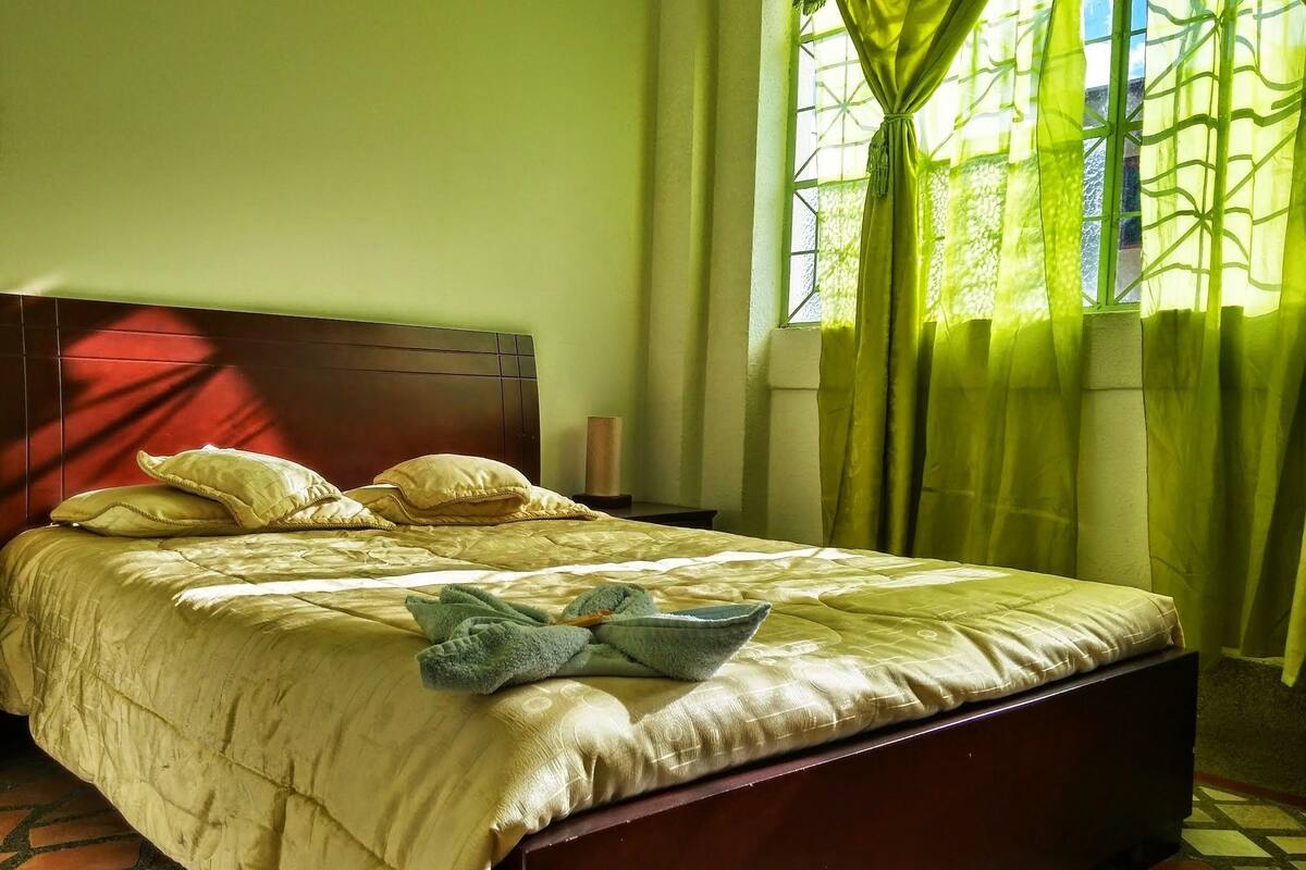 A neatly made bed with a headboard, a bedside table, and a window with green curtains.