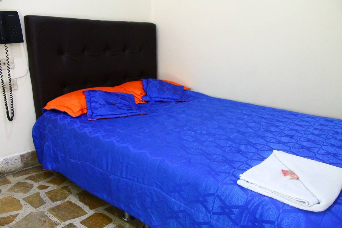 A single bed with a blue bedspread and orange pillows, a white towel on the bed, and a black telephone on the wall to the left.