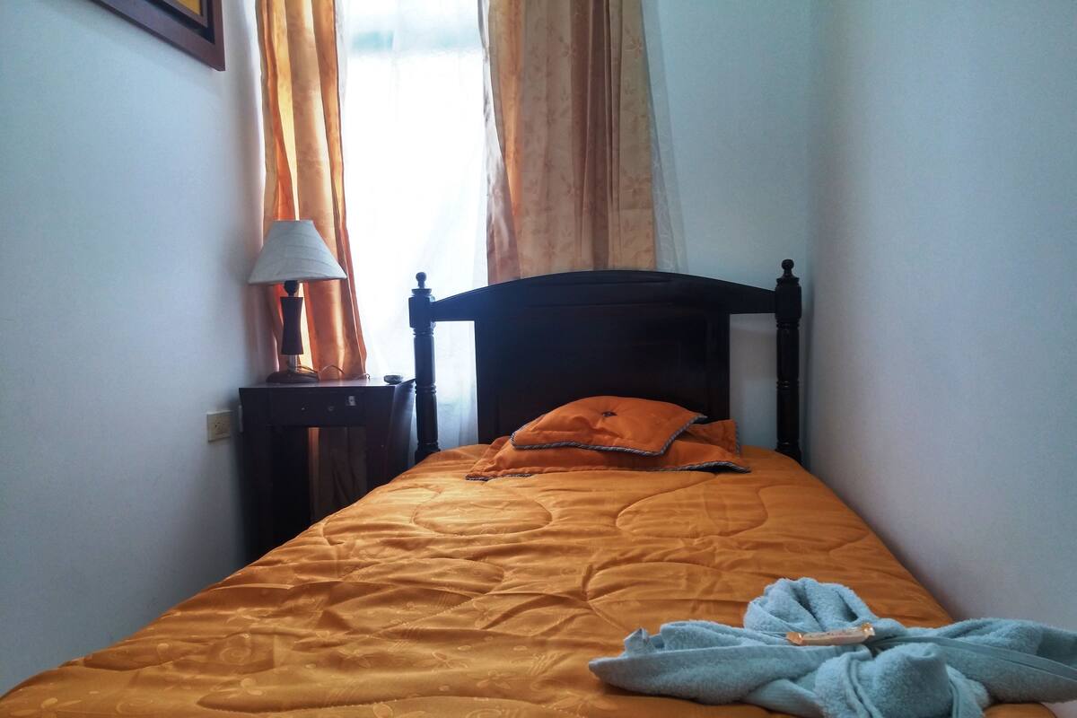 A neatly made bed with an orange bedspread and a blue towel, a wooden headboard, a bedside table with a lamp, and sheer curtains.
