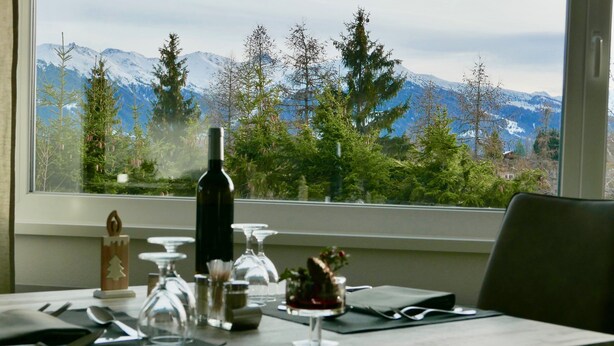 A dining table set with cutlery, a wine bottle, and a decorative item, with a mountain view through a window.