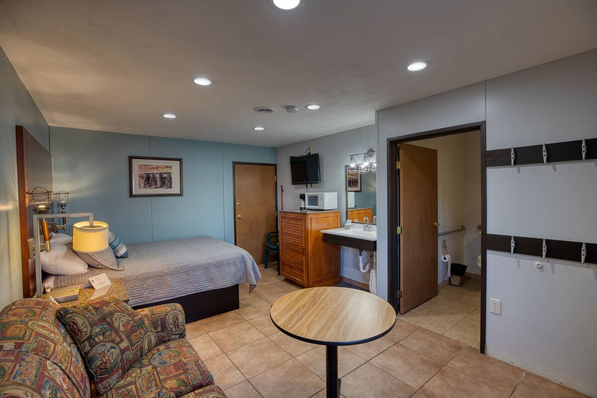 A bedroom with a bed, a sofa, a round table, a wardrobe, a dresser, a mirror, and a bathroom with a sink and toilet.