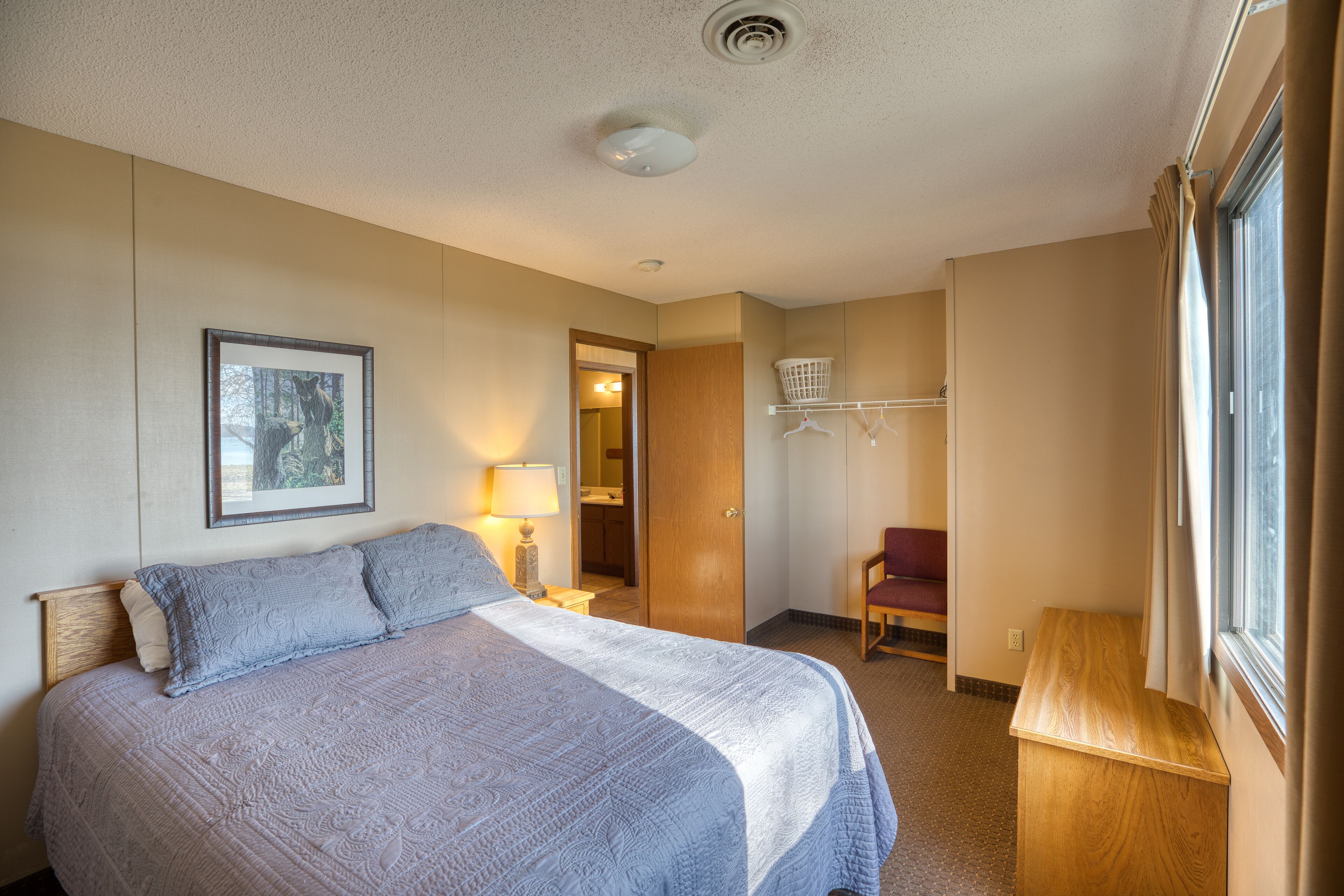 A hotel room with a large bed, a wooden nightstand, a chair, a window with curtains, a framed picture on the wall, and a door leading to a bathroom.