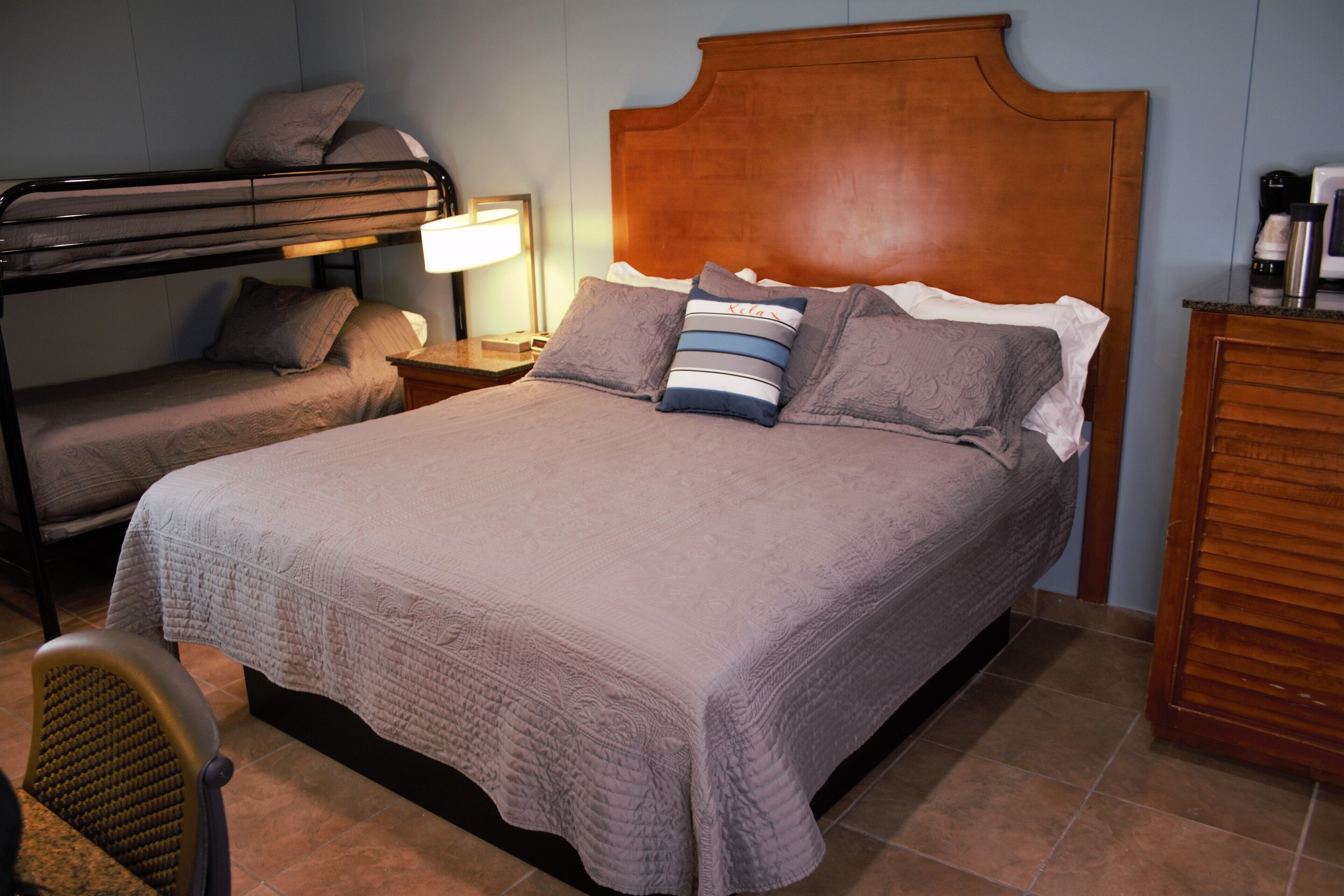 Cama doble con cabecera de madera, dos almohadas, mesita de noche con lámpara y una cama litera a la izquierda.