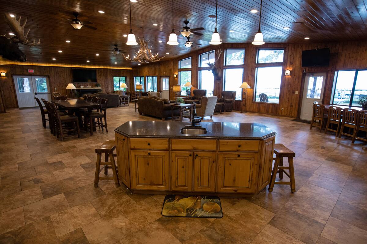 A spacious room with a large wooden dining table, multiple chairs, and a central island with a sink. There are ceiling fans, pendant lights, and large windows providing natural light.