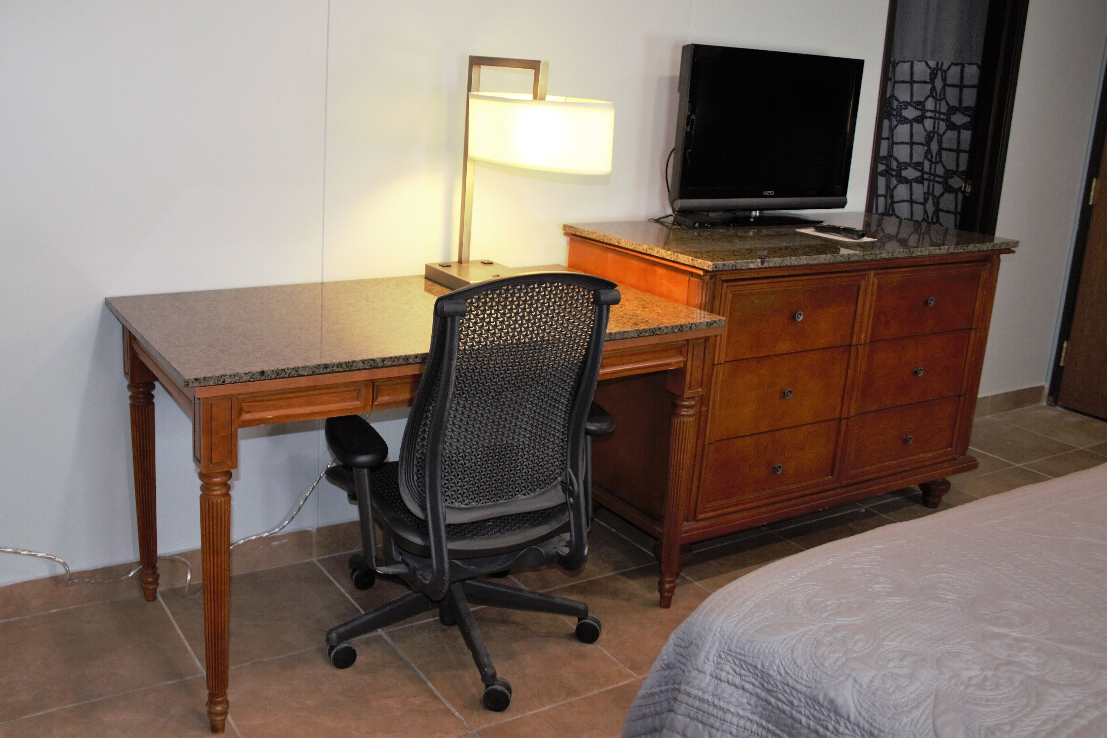 Una habitación de hotel con un escritorio de madera, una silla de oficina negra, un televisor sobre un aparador y una cama con edredón estampado.