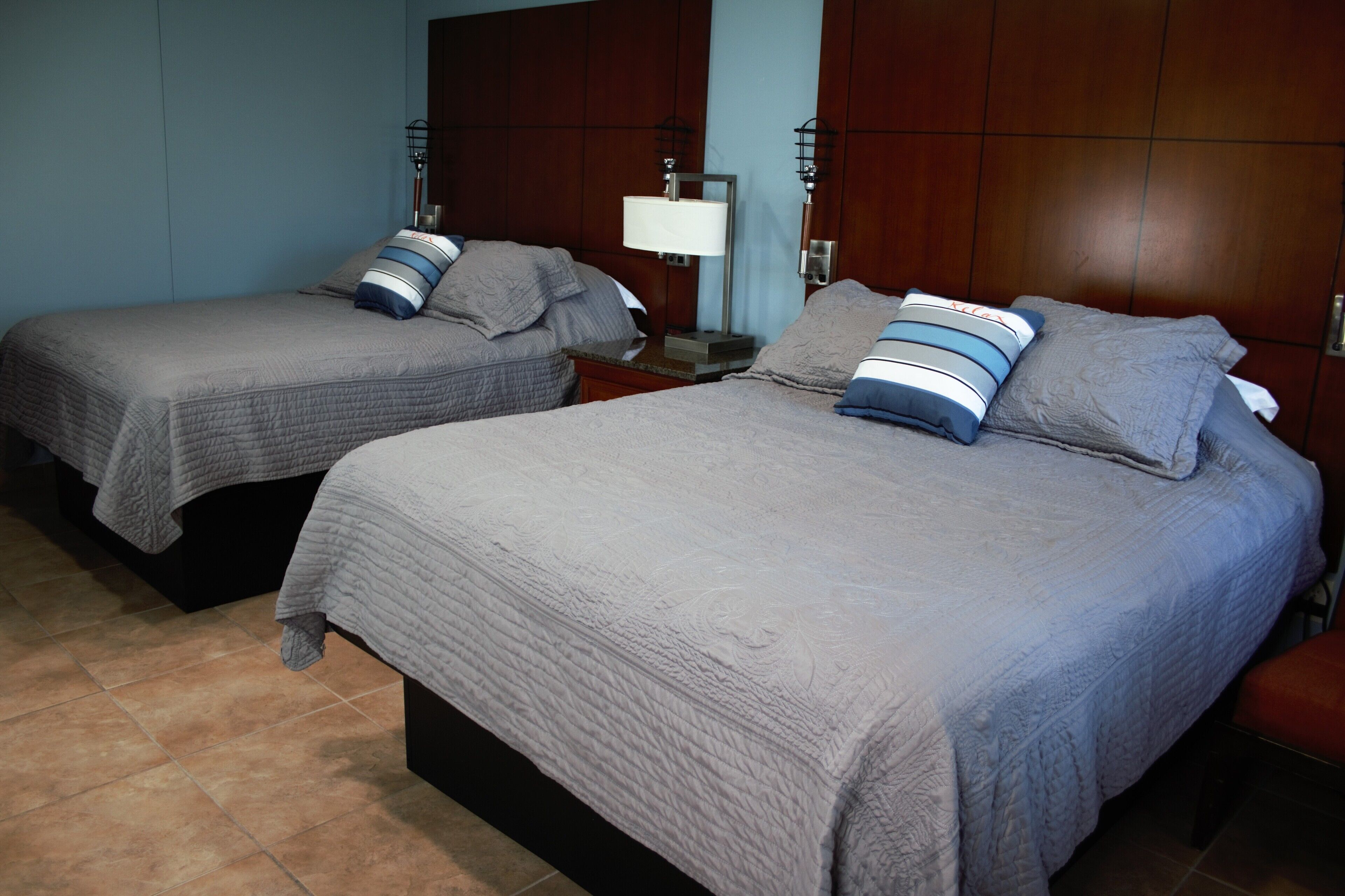 Dos camas individuales con ropa de cama gris y almohadas a rayas, una mesita de noche con una lámpara y un cabecero de madera.