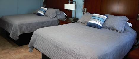 Dos camas individuales con ropa de cama gris y almohadas a rayas, una mesita de noche con una lámpara y un cabecero de madera.