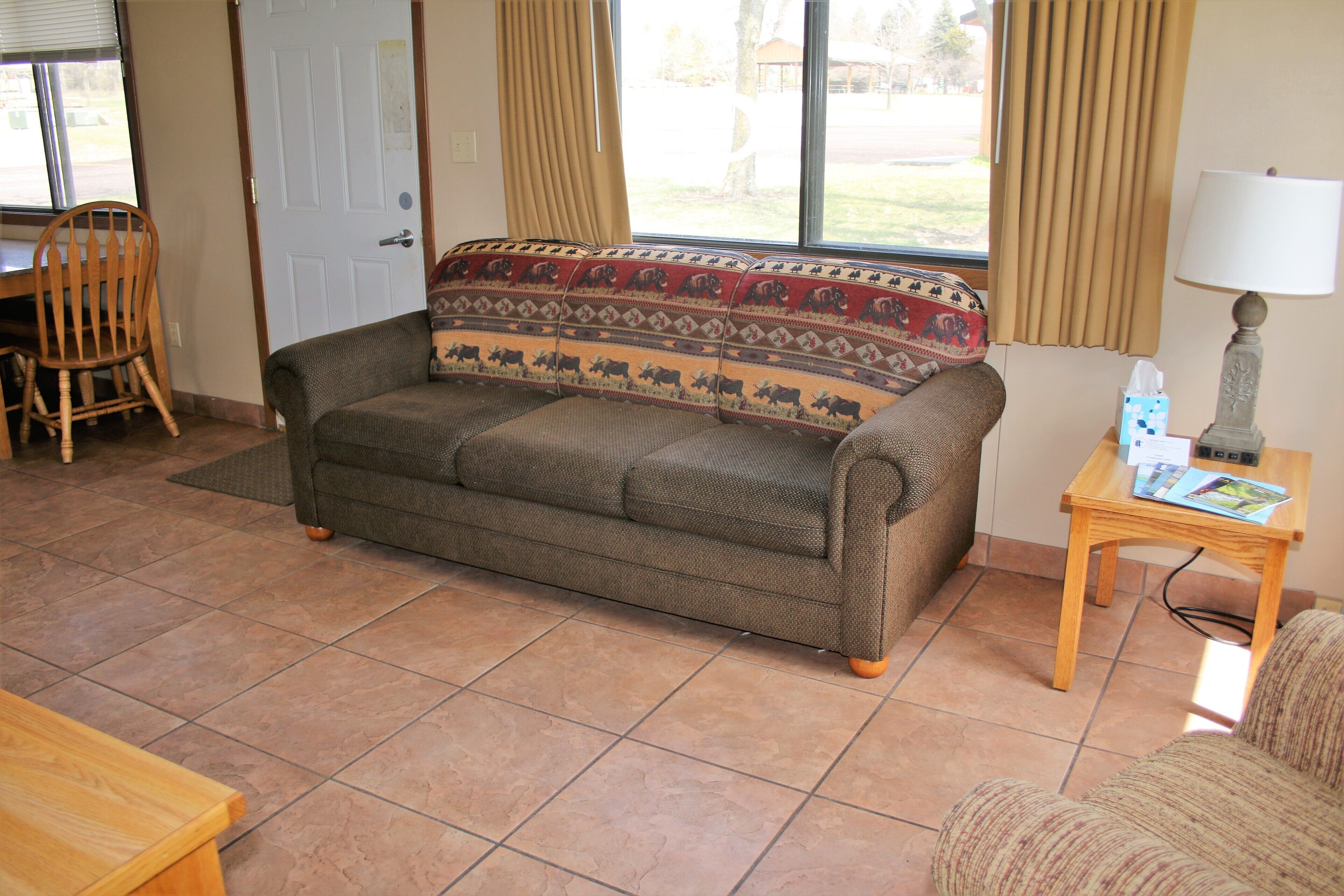 A living room with a sofa, a wooden chair, a small table with a lamp, and a window with curtains.