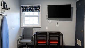 A room with a flat-screen TV mounted on the wall, a window with a geometric patterned curtain, a desk with two chairs, and a door with a vent. - Hillsboro Inn & Suites (Hillsboro)
