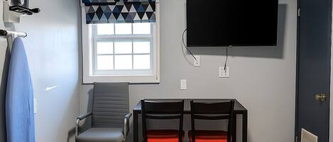 A room with a flat-screen TV mounted on the wall, a window with a geometric patterned curtain, a desk with two chairs, and a door with a vent.