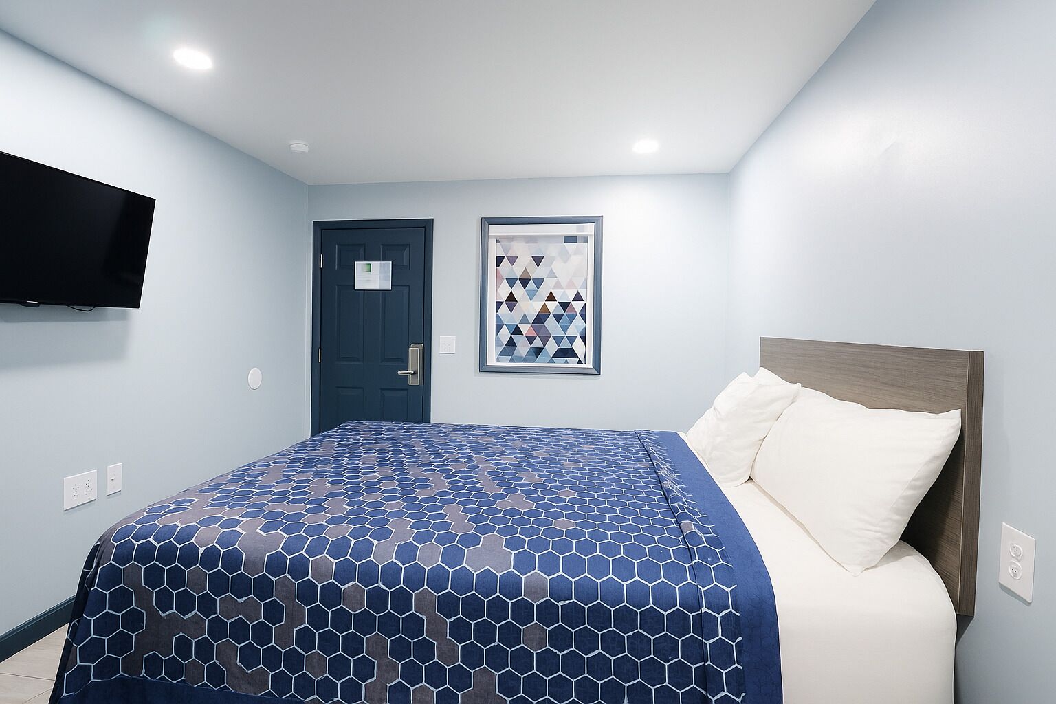 Una habitación de hotel con una cama, una puerta, un cuadro enmarcado y un televisor fijado a la pared.