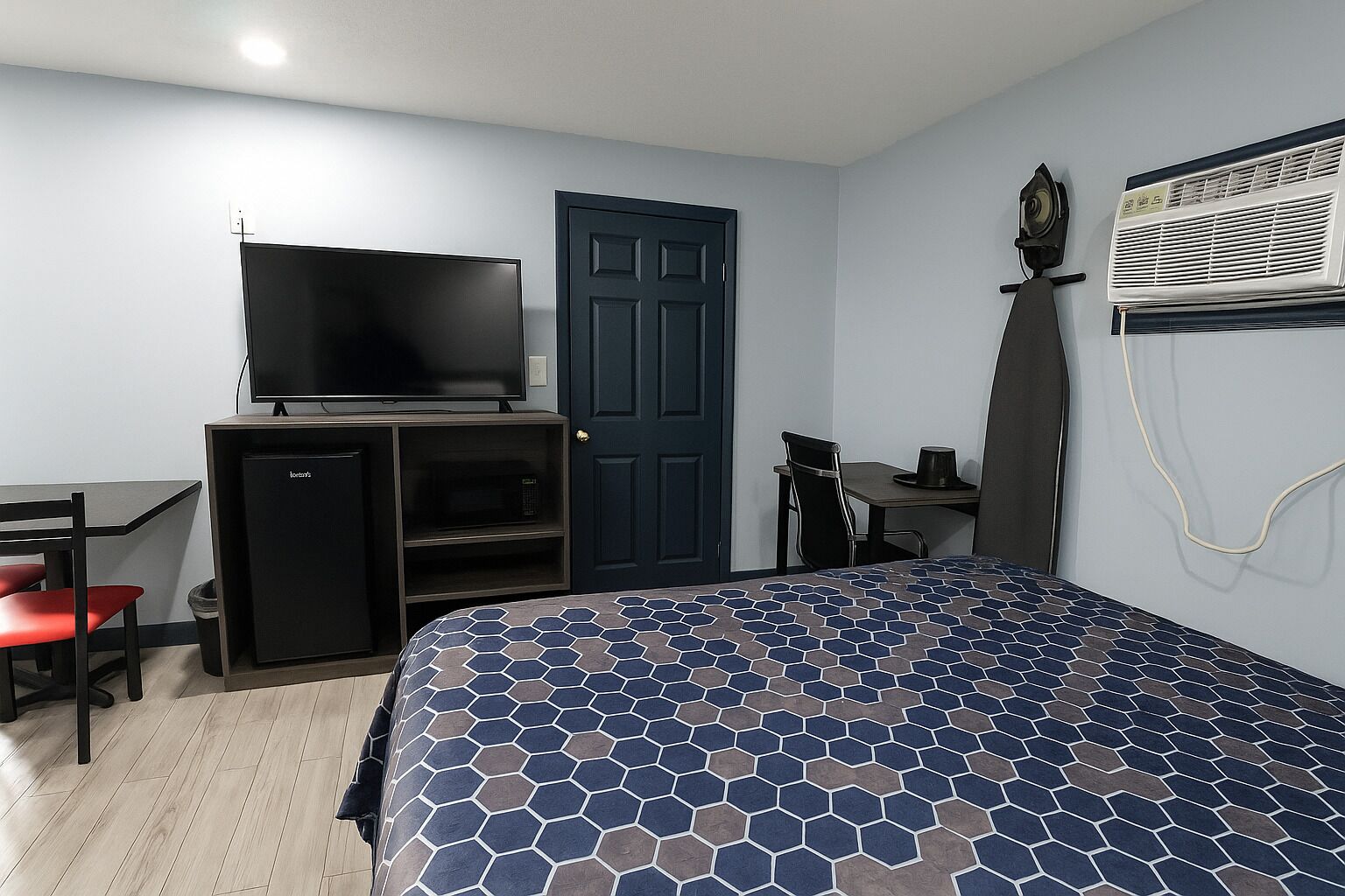 Habitación de hotel con cama, televisor sobre un mueble, escritorio con silla, puerta y equipo de aire acondicionado.