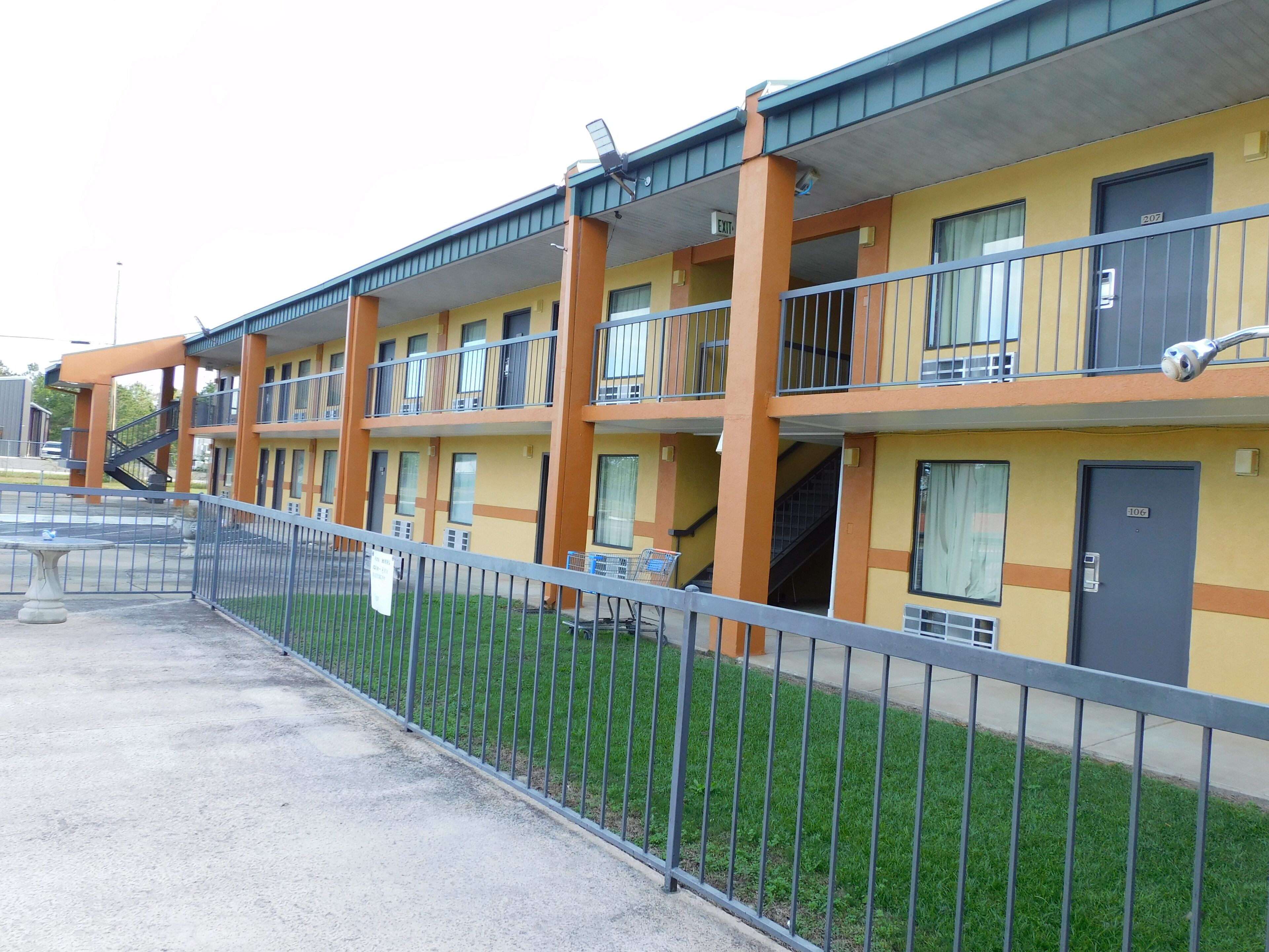 Ein Mehrstöckiges Hotel mit Balkonen, einem umzäunten Parkplatz und einem Rasen.