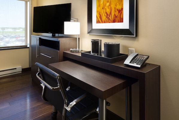 Une chambre d’hôtel avec un bureau en bois, un fauteuil de bureau noir, un téléviseur à écran plat, une œuvre d’art encadrée, un téléphone et une fenêtre donnant sur l’extérieur.