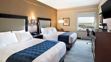 A hotel room with two beds, a desk, a chair, a TV, and a window with a view of an airport.