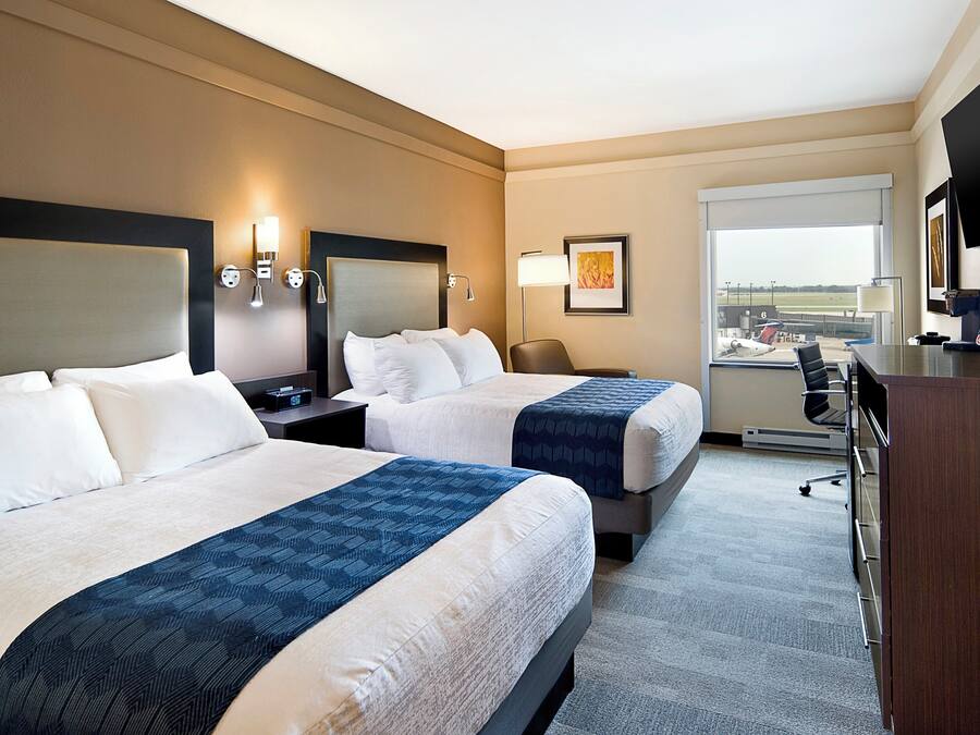 A hotel room with two beds, a desk, a chair, a TV, and a window with a view of an airport.