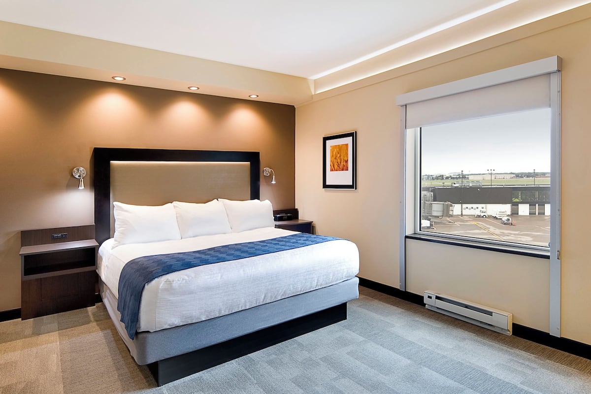 A modern hotel room with a large bed, a nightstand, a wall-mounted lamp, a framed picture, and a window with a view of an airport.