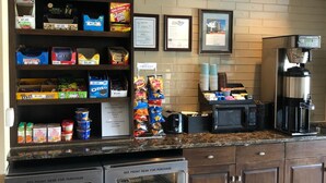 Uma cozinha compacta em hotel com a marca AeroStay, prateleiras para lanches, uma cafeteira e um refrigerador.