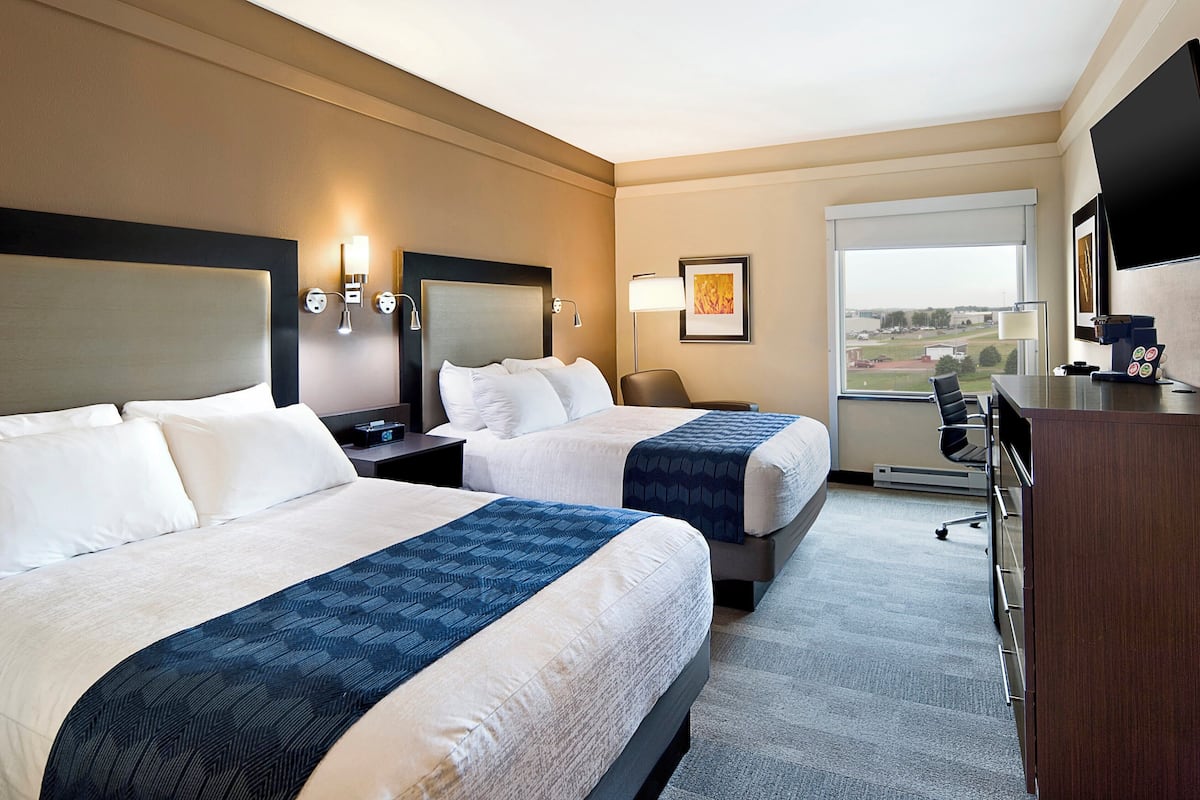 A hotel room with two beds, a desk, a chair, a TV, and a large window.