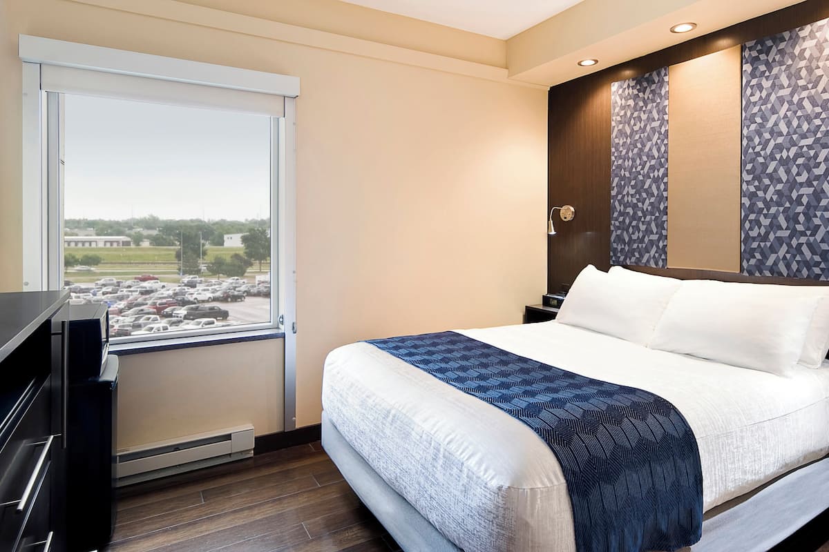 A hotel room with a bed, a TV, a window with a view of a parking lot, and a wall with a geometric pattern.