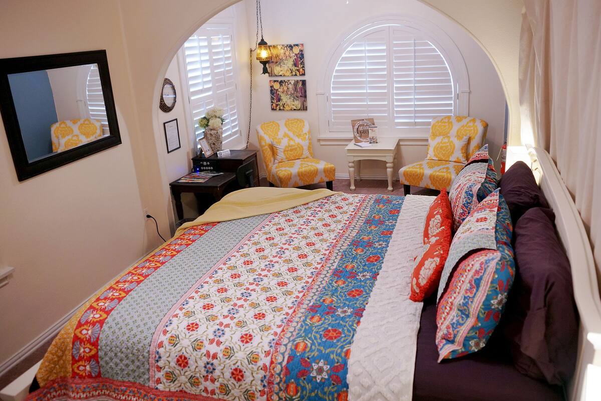A bedroom with a large bed, two yellow armchairs, a small table, and a window with blinds.