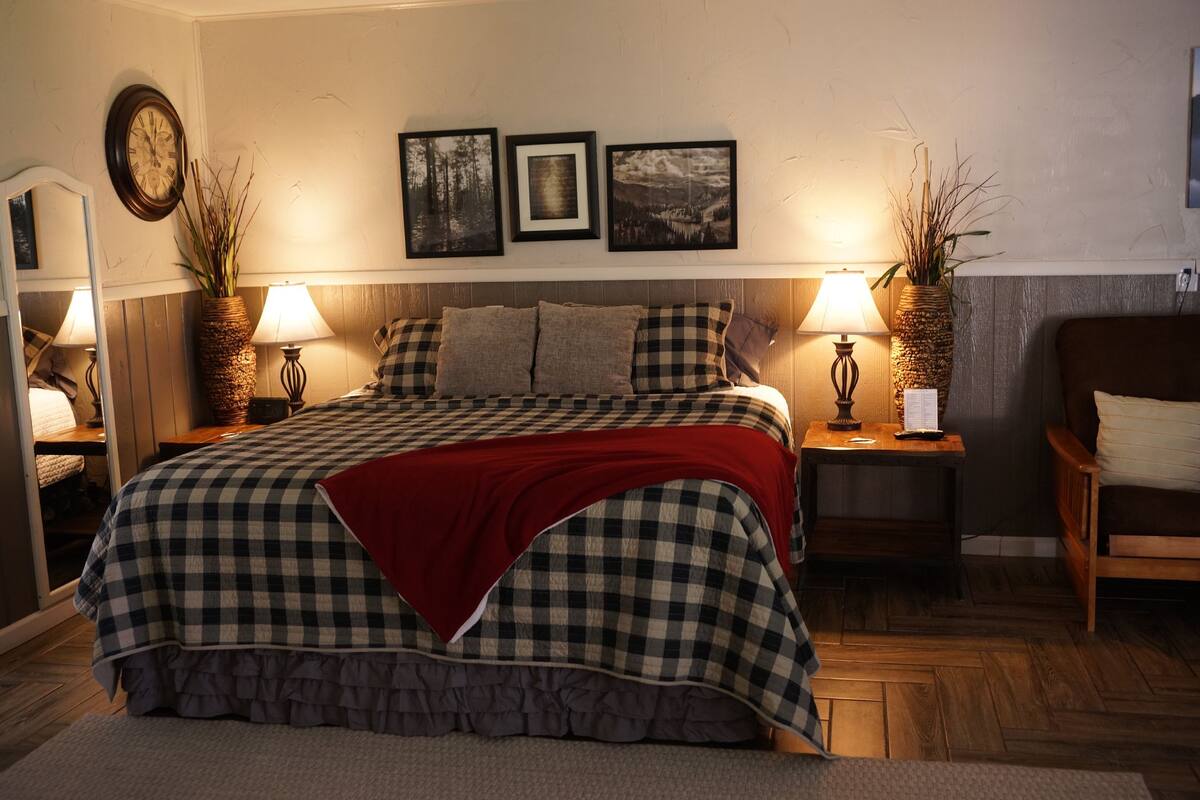 A neatly arranged bedroom with a checkered bedspread, a red throw blanket, two bedside tables with lamps, a clock on the wall, and three framed pictures above the bed.