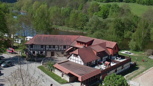 A traditional building with a red roof and white walls, situated near a lake with a parking area in front. - Landgasthof Probstei Zella (Amt Creuzburg)