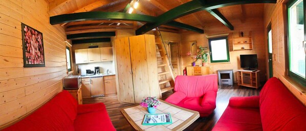 Interior de una cabaña de madera con cocina, comedor y sala de estar, con mobiliario rojo y vigas de madera.