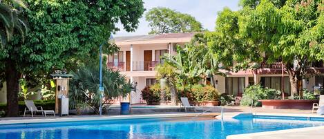 A swimming pool with lounge chairs, surrounded by lush greenery and a multi-story building with balconies.