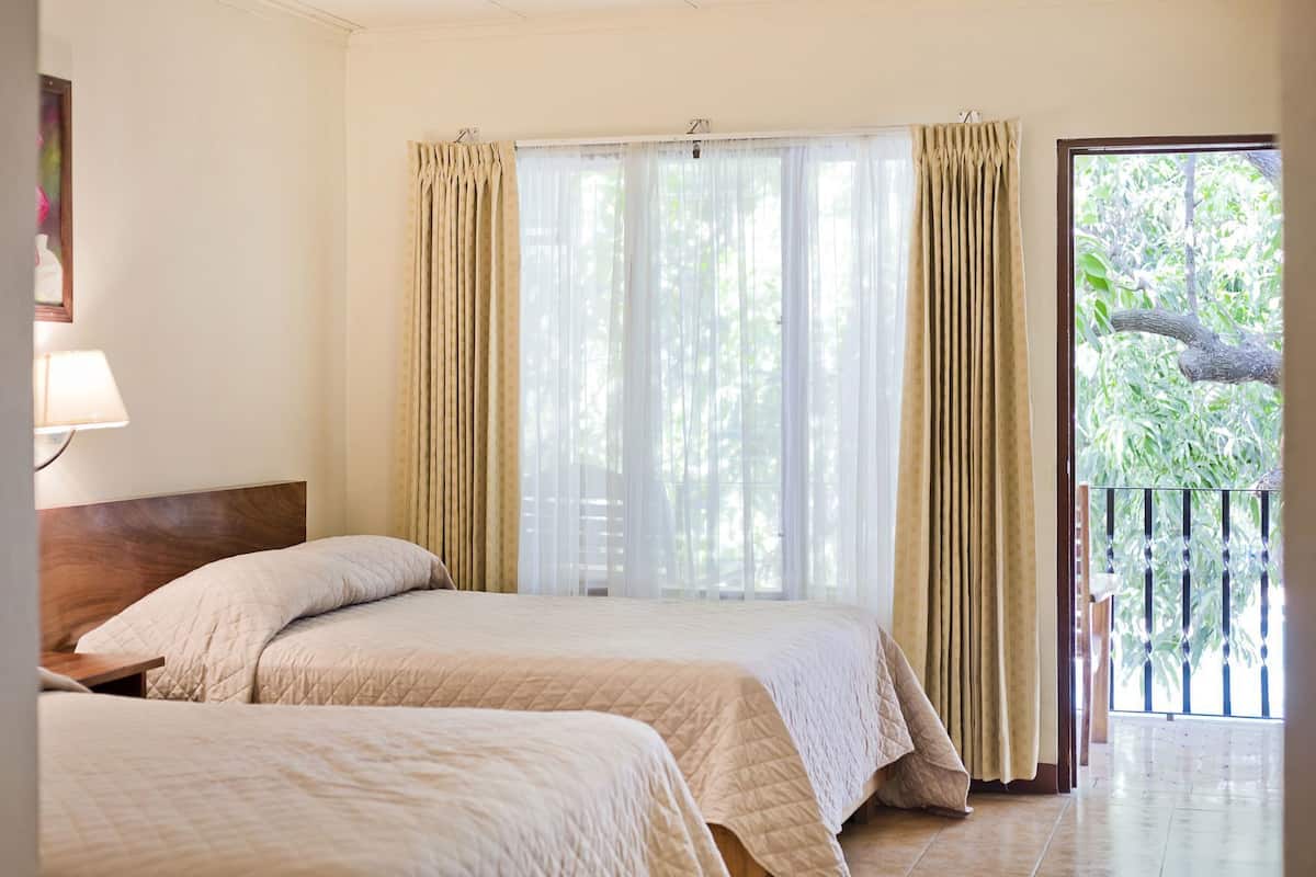 Une chambre avec deux lits, une fenêtre ornée de rideaux légers, une vue sur la verdure et un balcon avec une balustrade.