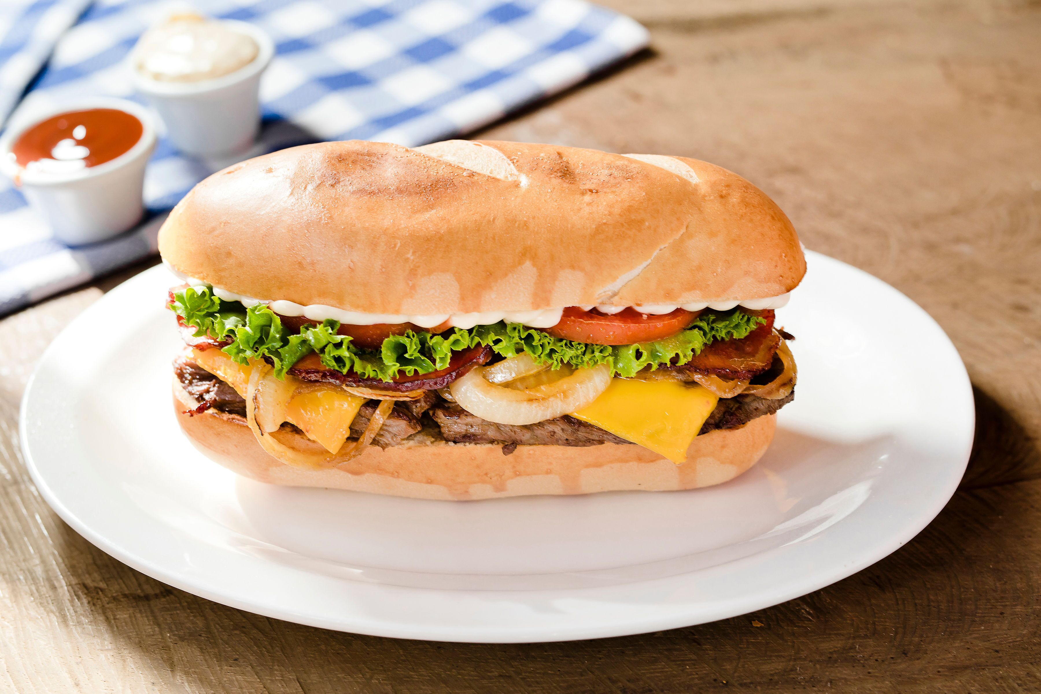 Un sándwich sobre un plato blanco, con una servilleta azul y blanca a cuadros y pequeños recipientes con salsas al fondo.