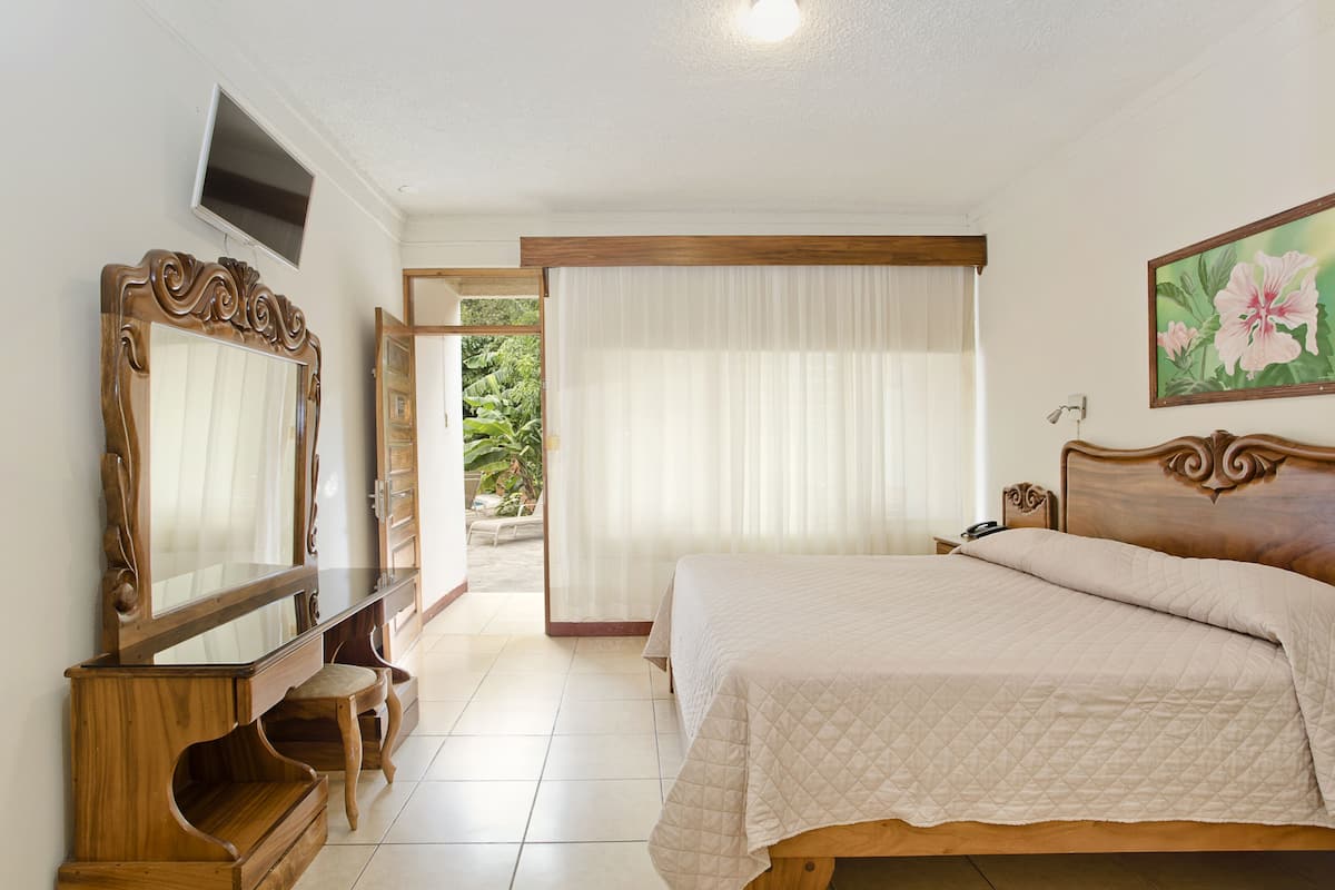 Une chambre à coucher avec un lit, une commode en bois, un téléviseur et une fenêtre avec des rideaux.