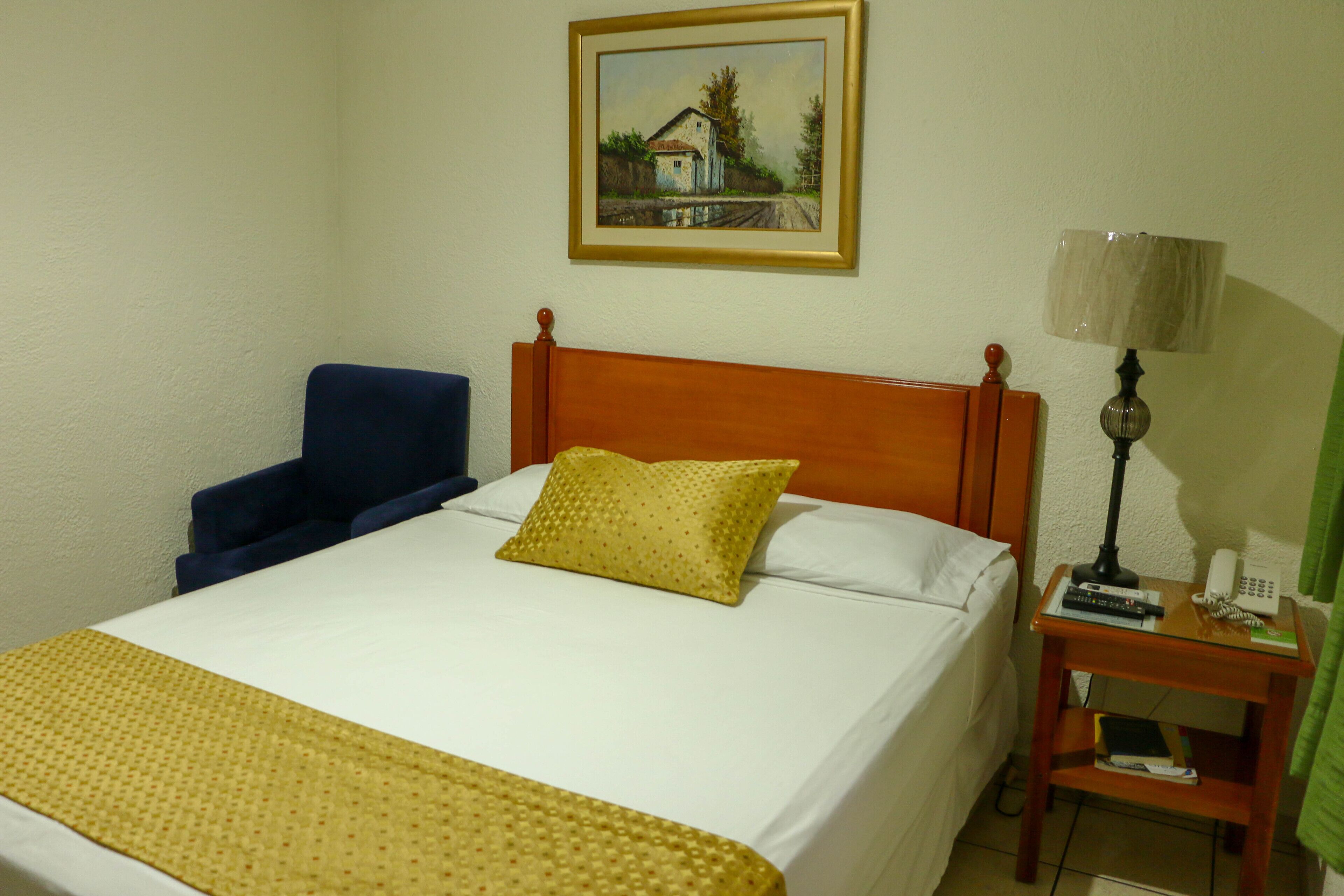 A hotel room with a single bed, a chair, a bedside table with a lamp, and a painting on the wall.