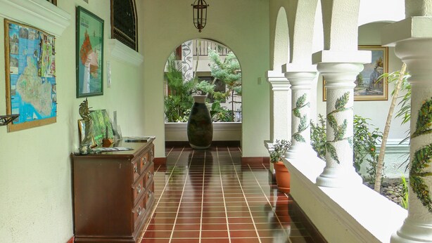 Um corredor com piso de azulejos, vigas de madeira no teto e colunas brancas com arcos.