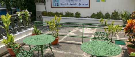 A patio area with green metal tables and chairs, surrounded by potted plants and a sign for 'Hoteles Villa Serena'.