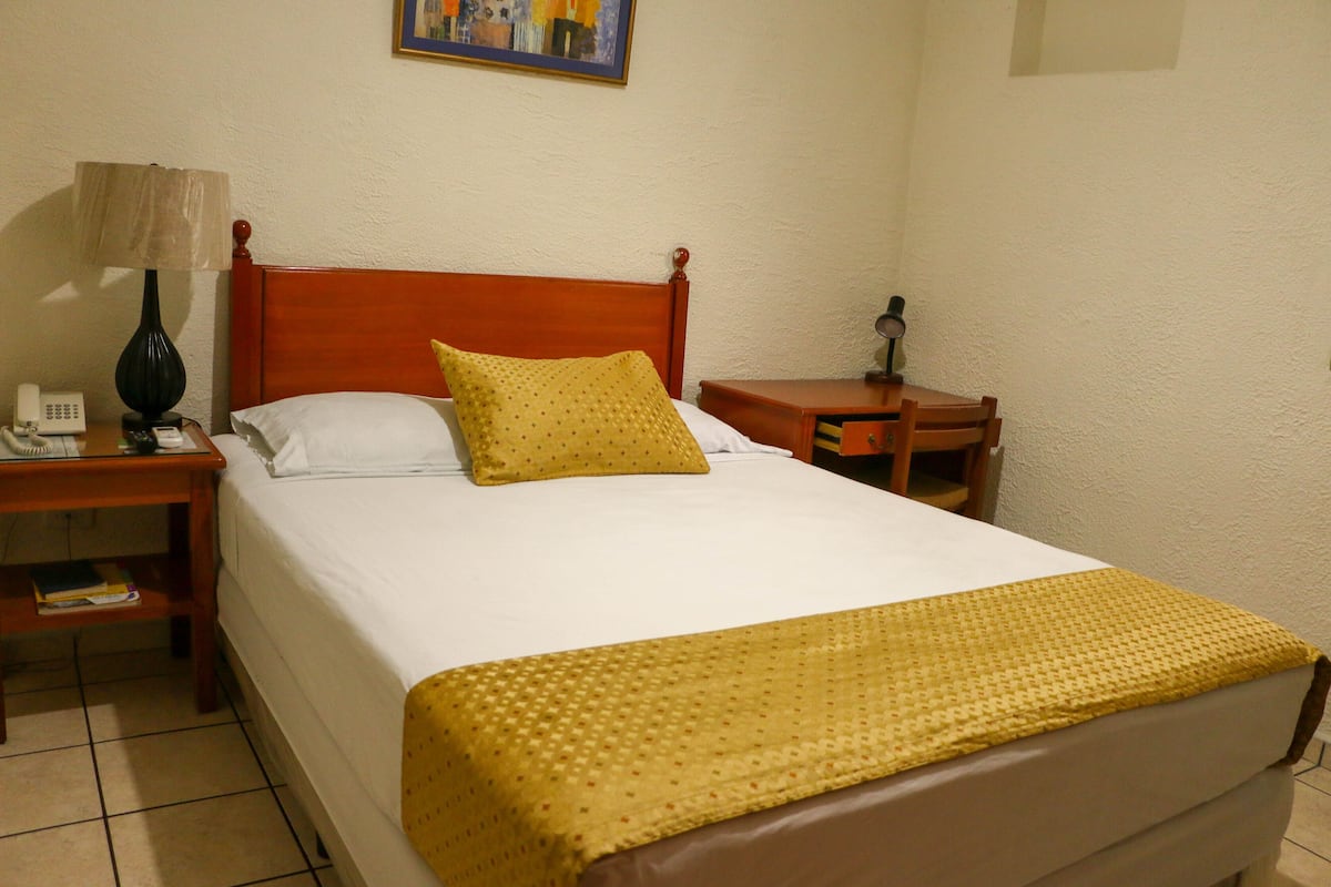 Ein Hotelzimmer mit einem hölzernen Bett, einem Nachttisch mit Lampe, einem Beistelltischchen mit Telefon und einem kleinen Tisch mit Lampe.