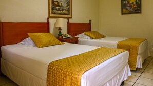 Quarto de hotel com duas camas, cada uma com roupa de cama branca e cabeceiras amarelas. Há um quadro na parede acima das camas e uma luminária na mesa de cabeceira entre elas.