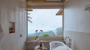A bathroom with a freestanding bathtub, a window with a view of greenery, and a shelf with toiletries.