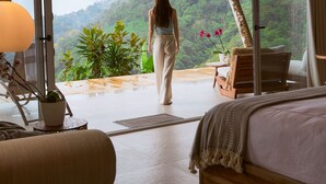 Une pièce spacieuse avec un grand lit, un coin salon comprenant un fauteuil et une table, et une vue sur une forêt luxuriante grâce à de grandes fenêtres.